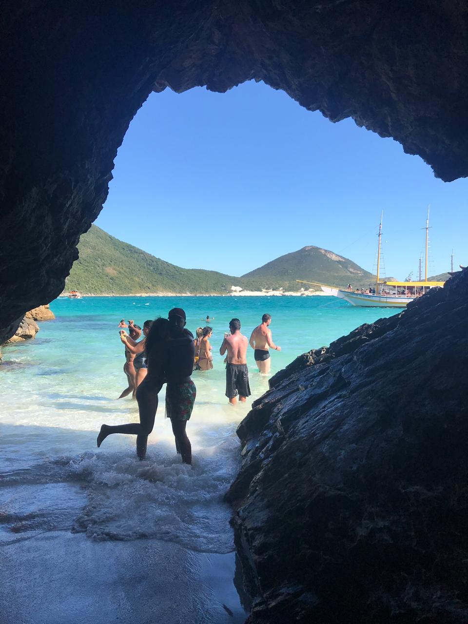 Casal abraçado posando pra foto com a gruta formando uma moldura na foto, mar e ilha do farol ao fundo